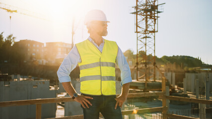 Confident Bearded Head Civil Engineer-Architect in Sunglasses Stands in a Construction Site on a Sunny Bright Day. Man is Wearing a Hard Hat, Shirt, Jeans and a Yellow Safety Vest. 