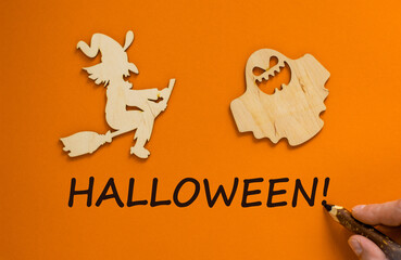 Hand with black pencil writing 'halloween'. Wooden witch and ghost. Beautiful orange background. Celebrate concept.