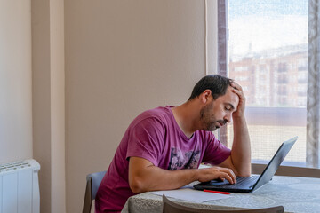 Bearded man wearing purple t-shirt looking at bills worried in his living room at home