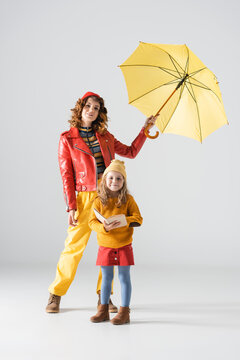 Mother And Daughter In Colorful Red And Yellow Outfits With Umbrella And Book On Grey Background