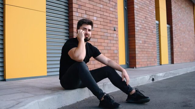 Happy young man talking on the phone enthusiastically while sitting on the sidewalk in the middle of the city. Successful businessman dressed modestly talking on the phone happily.