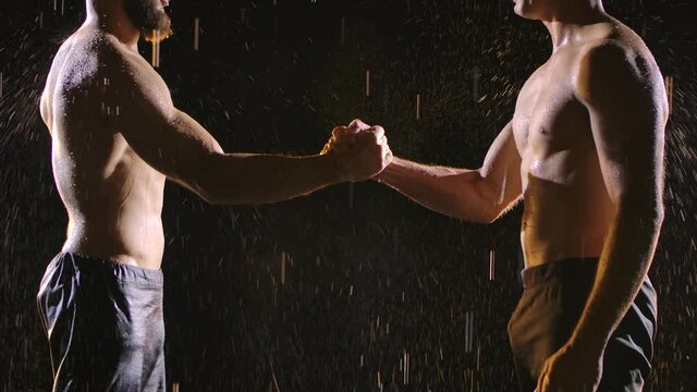 Two muscular topless men shake hands in the splashing rain. Picture taken in the studio on a black background. Slow motion. Close up.