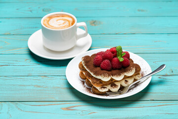 portion of Classic tiramisu dessert with raspberries and cup of espresso coffee on blue wooden background