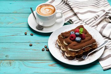 portion of Classic tiramisu dessert with raspberries and blueberries and cup of espresso coffee on blue wooden background