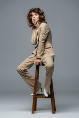 elegant woman in beige suit posing on wooden chair on grey background