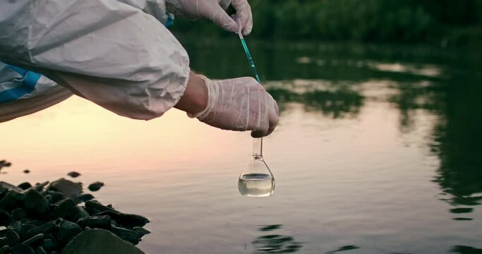 Male ecologist's hands in rubber gloves taking water probes from dirty polluted river. Chemist working, holding glass tube with chemical agent and pipette, analysing level of environmental pollution.