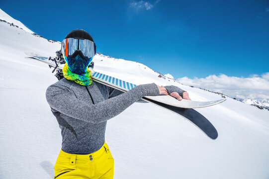 A sporty slender girl without a jacket with a closed face scarf and wearing a ski mask with goggles stands with skis on her shoulders