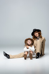 elegant mother and daughter in white and beige outfits and hats posing on floor on grey background