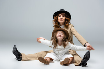 elegant mother and daughter in white and beige outfits and hats posing on floor on grey background