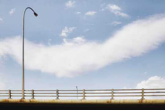 A street light curving over a highway bridge with a guard rail looked at from the bottom up on a cloudy summer day
