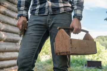 Male carpenter holds a homemade wooden box in one hand and a hammer in the other, against the backdrop of an old log barn.
