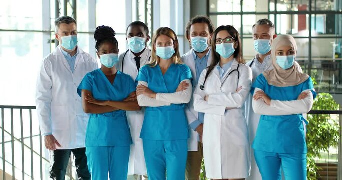 Mixed-races Busy Team Of Male And Female Doctors Posing To Camera And Crossing Hands In Hospital. International Group Of Medics In Medical Masks. Protected Multi Ethnic Physicians And Nurses In Clinic