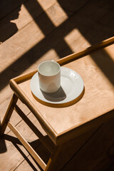 white cup of coffee on a white saucer on wooden tray in morning lights