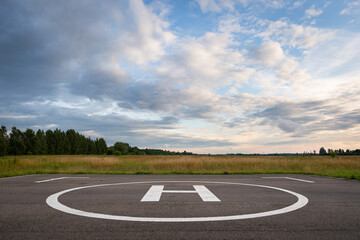 A closeup of an asphalt-covered helipad with a special symbol in the center for helicopter landing, against the backdrop of a green field and a cloudy evening sky.