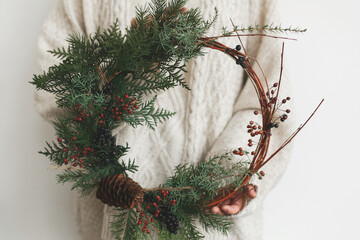 Woman in stylish knitted sweater holding modern christmas wreath on white background