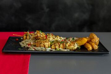 Shish kebab from chicken breast on bamboo sticks, baked potatoes sprinkled with green onions with original serving on a black plate.