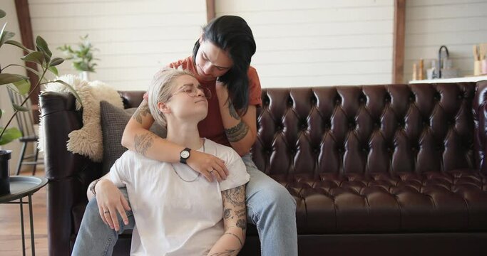 Joyful lesbian couple with various tattoos of black colour hugs and kisses sitting at home against white wall slow motion