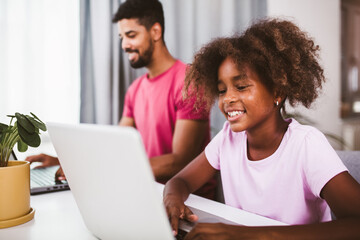 Father using laptop while his daughter is doing homework in the living room.