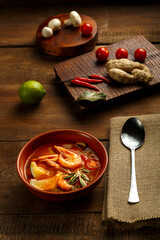 Tom yam soup with shrimp and lime in a brown dish on a wooden table next to a spoon on a napkin and ingredients lime ginger.