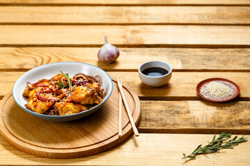 plate of soba with chicken and sesame seeds in teriyaki sauce on a wooden stand and sticks next to sesame seeds and soy sauce on a wooden table.