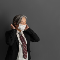 Elegant mature businesswoman putting on protective mask. Serious female office worker on gray back. Coronavirus pandemic concept.