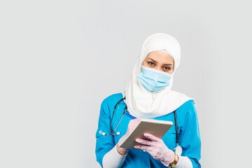 friendly muslim doctor or nurse wearing hijab and medical face mask with stethoscope writing prescription to patient on gray background