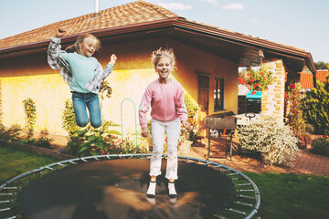 Happy two blonde girls are jumping on a trampoline in the garden near their house. Leisure and games - obrazy, fototapety, plakaty
