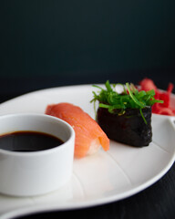 white plate in the shape of a leaf on which there is soy sauce, sushi with salmon, green seaweed and red ginger on a dark stone background
