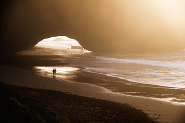 Legzira beach, Morocco, Africa. Silhouette of a pair, man and woman walk along the shore, Atlantic ocean, wet sand and sea surf at the background of famous mountain arch in the light of setting sun