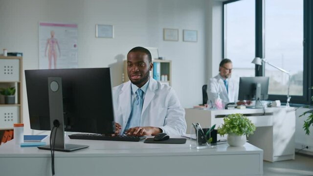 Happy Smiling African American Medical Doctor is Working on a Computer in a Health Clinic. Physician in White Lab Coat is Browsing Medical History Behind a Desk in Hospital Office. 