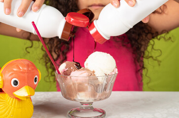 Happy child putting syrup in a bowl of ice cream on a table with green background, selective focus.