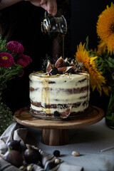 Woman pouring syrup on cake top with fresh figs, blackberries and pistachios. Layered sponge cake with cream cheese on wooden stand surrounded with flowers on black background.