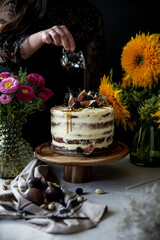 Woman pouring syrup on cake top with fresh figs, blackberries and pistachios. Layered sponge cake with cream cheese on wooden stand surrounded with flowers on black background.