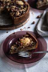 Piece of frangipani tart with pears, hazelnuts and chocolate on glass purple plate on linen tablecloth .