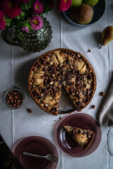 Top view of frangipani tart with pears, hazelnuts and chocolate, plates, fresh pears and vase with flowers on linen tablecloth .