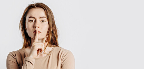 Shh gesture. Young beautiful serious girl holding a finger to her lips on a white isolated background. A woman asks to be silent, a place for advertising. Negative brunette in a beige jumper.