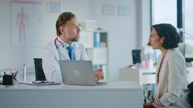 Family Doctor is Delivering Great News About Female Patient's Medical Results During Consultation in a Health Clinic. Physician in White Lab Coat Sitting Behind a Computer in Hospital Office. 