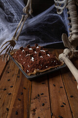 Kuchen als Friedhof mit Gräbern und Skeletten aus Schokokrümel, Creme und Banane auf Tisch aus Holz und dahinter Spinnweben