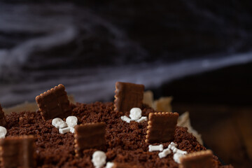 Kuchen als Friedhof mit Gräbern und Skeletten aus Schokokrümel, Creme und Banane auf Tisch aus Holz und dahinter Spinnweben
