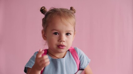 childhood, school, education, upbringing, science concept - close-up little blonde caucasian slavic girl in blue dress with backpack holds book and sternly waves index finger on solid pink background
