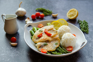 Fried tilapia, asparagus, cherry tomatoes, lemon and rice balls on a white plate. 