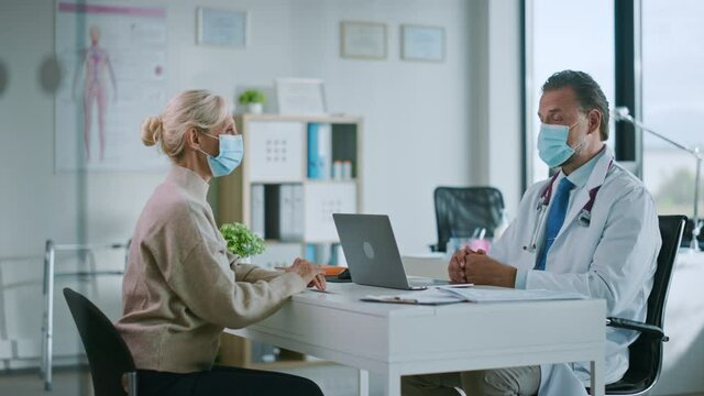 Family Doctor in Protective Mask is Reading Medical History of Senior Female Patient and Speaking with Her During Consultation in a Health Clinic. Physician in Lab in Front of Computer in Hospital.
