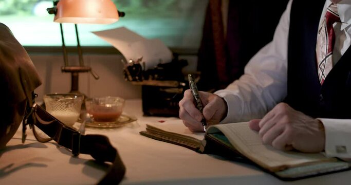 A businessman from the 1940's makes notes in his ledger as he sits by a window of the dinning car of a passenger train he is traveling on.