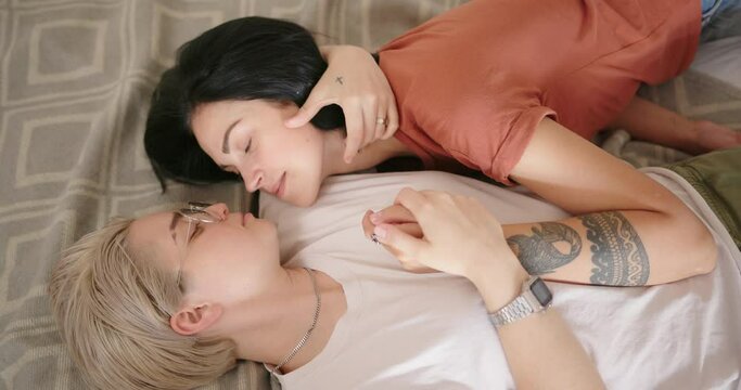 Romantic lesbian couple in coloured t-shirts lies on brown blanket and kisses joining hands at home slow motion close view