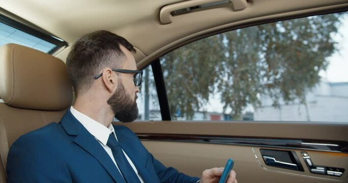 Young handsome Caucasian man in suit, tie and glases sitting on backseat of expensive car and holding cellphone. Serious businessman with beard and mobile phone, going somewhere and looking at window.