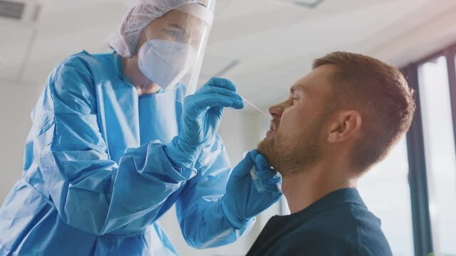Medical Nurse in Safety Gloves and Mask, Protective Face Shield and Overalls is Taking a PCR Corona Virus Sample in a Health Clinic. Doctor Uses Respiratory Swab Test. Covid-19 Pandemic Concept.