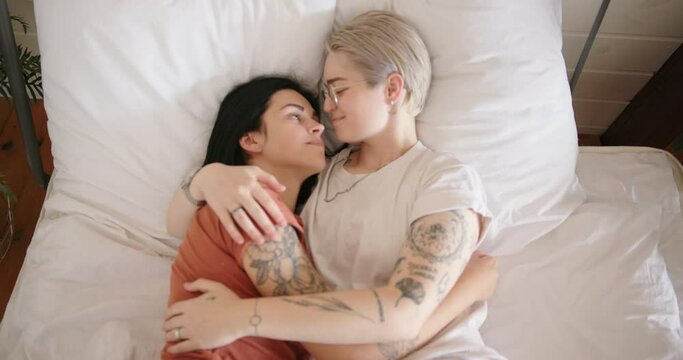 Smiling young woman in white t-shirt and glasses hugs tattooed girlfriend lying on bed between green pot plants slow motion