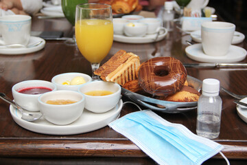 new normal in restaurants, table view with breakfast foods, sweet bread, coffee, juice and natural jams with protection mask and antibacterial gel