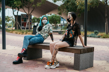 Chicas modernas hablando sobre la actualidad en un banco al aire libre con mascarillasy meidas de...