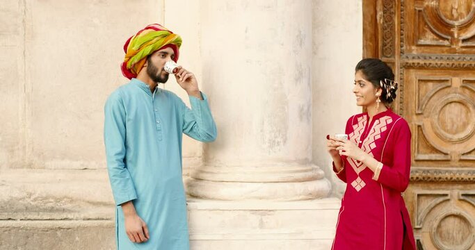 Hindu happy woman with dot on forehead and man in turban standing outdoors, talking and drinking coffe or tea. Resting. Handsome male and beautiful female sipping drink and spending time together.